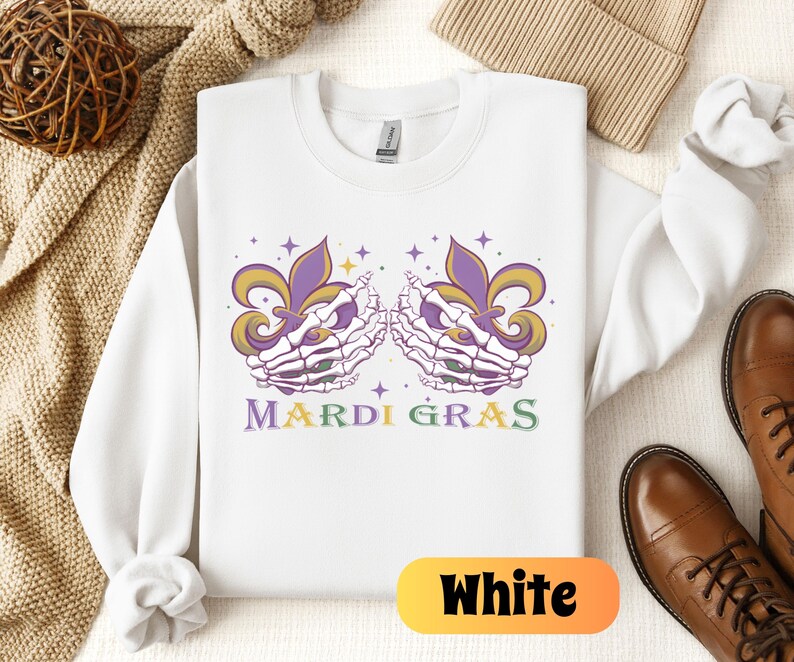 May include: White sweatshirt with a purple and green Mardi Gras design featuring two skeletal hands holding fleur-de-lis. The text "Mardi Gras" is printed in green and yellow.