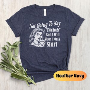 May include: A navy blue t-shirt with a retro-style illustration of a woman holding a cup of coffee. The text on the shirt reads "Not Going To Say "I Told You So" But I Will Wear It On A Shirt".