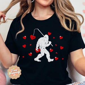 May include: Black t-shirt with a white graphic of a Bigfoot holding a fishing rod and catching hearts. The graphic is surrounded by red hearts.
