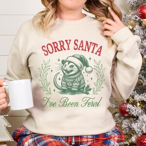 May include: A cream-colored sweatshirt with the text "Sorry Santa I've Been Feral" in red and green. A cartoon possum wearing a Santa hat and a green and red sweater is in the center of the design.