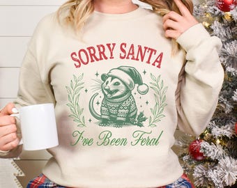 May include: A cream-colored sweatshirt with the text "Sorry Santa I've Been Feral" in red and green. A cartoon possum wearing a Santa hat and a green and red sweater is in the center of the design.