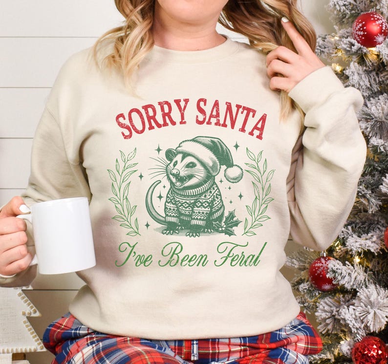 May include: A cream-colored sweatshirt with the text "Sorry Santa I've Been Feral" in red and green. A cartoon possum wearing a Santa hat and a green and red sweater is in the center of the design.