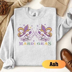 May include: A gray sweatshirt with a Mardi Gras design featuring two skeletal hands holding purple, green, and gold fleur-de-lis. The text "Mardi Gras" is printed below the hands.