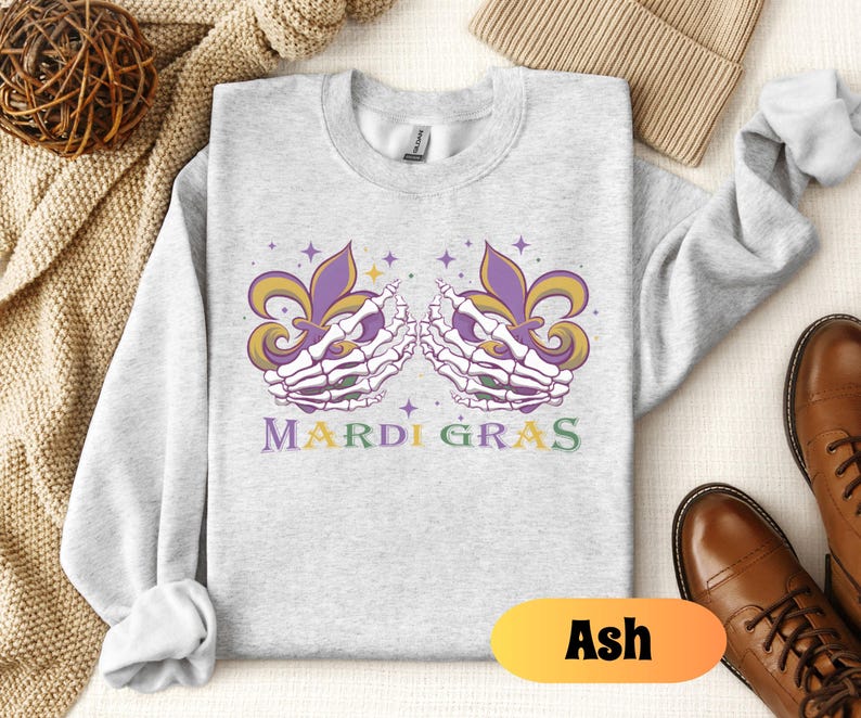 May include: A gray sweatshirt with a Mardi Gras design featuring two skeletal hands holding purple, green, and gold fleur-de-lis. The text "Mardi Gras" is printed below the hands.