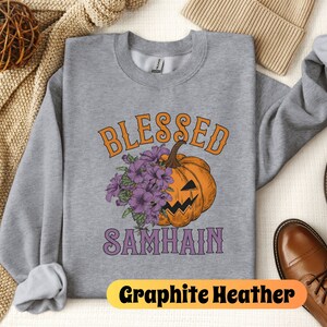 以下が含まれることがあります： グレーのトレーナーに、紫とオレンジ色の「Blessed Samhain」という文字が書かれています。文字の中央には、紫の花束を持ったアニメーションの南瓜が描かれています。トレーナーは、茶色の帽子と茶色の靴を置いた白い表面の上に置かれています。