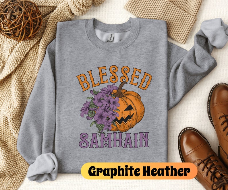 以下が含まれることがあります： グレーのトレーナーに、紫とオレンジ色の「Blessed Samhain」という文字が書かれています。文字の中央には、紫の花束を持ったアニメーションの南瓜が描かれています。トレーナーは、茶色の帽子と茶色の靴を置いた白い表面の上に置かれています。