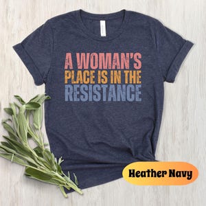 May include: Heather navy t-shirt with the text "A Woman's Place Is In The Resistance" in pink, orange, and blue. The shirt is displayed on a light wood surface with a sprig of green leaves.