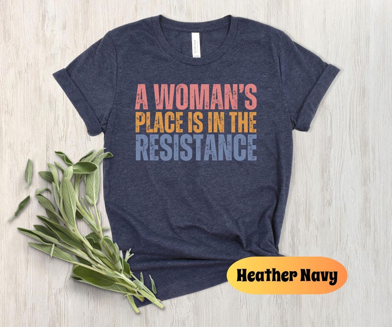 May include: Heather navy t-shirt with the text "A Woman's Place Is In The Resistance" in pink, orange, and blue. The shirt is displayed on a light wood surface with a sprig of green leaves.