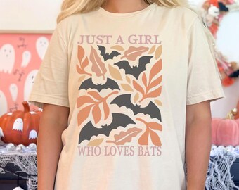 May include: A beige t-shirt with a graphic print of bats and leaves. The text "Just a girl who loves bats" is printed on the shirt.