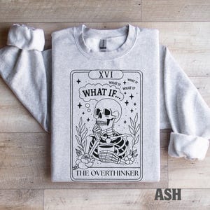 Puede incluir: Sudadera gris claro con un dise&ntilde;o de carta del tarot. El dise&ntilde;o presenta un esqueleto con una taza, el texto "WHAT IF..." y "THE OVERTHINKER". Las mangas est&aacute;n dobladas. La palabra "ASH" est&aacute; impresa en la parte inferior.