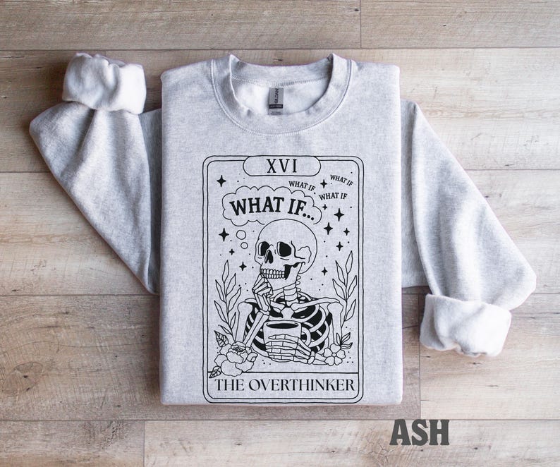 Puede incluir: Sudadera gris claro con un dise&ntilde;o de carta del tarot. El dise&ntilde;o presenta un esqueleto con una taza, el texto "WHAT IF..." y "THE OVERTHINKER". Las mangas est&aacute;n dobladas. La palabra "ASH" est&aacute; impresa en la parte inferior.