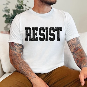 May include: White t-shirt featuring the word "RESIST" in large, distressed black lettering. The top has a crew neck design.