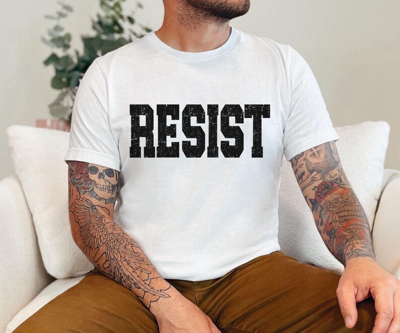 May include: White t-shirt featuring the word "RESIST" in large, distressed black lettering. The top has a crew neck design.