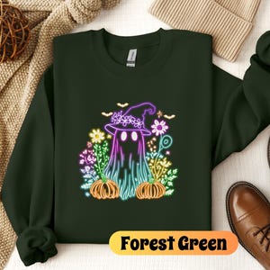 K&ouml;nnte beinhalten: Waldgr&uuml;ner Sweatshirt mit einem neongr&uuml;nen, violetten und rosafarbenen Geist, der einen Hexenhut tr&auml;gt und von Blumen und K&uuml;rbissen umgeben ist. Der Text "Forest Green" ist unten auf dem Bild aufgedruckt.