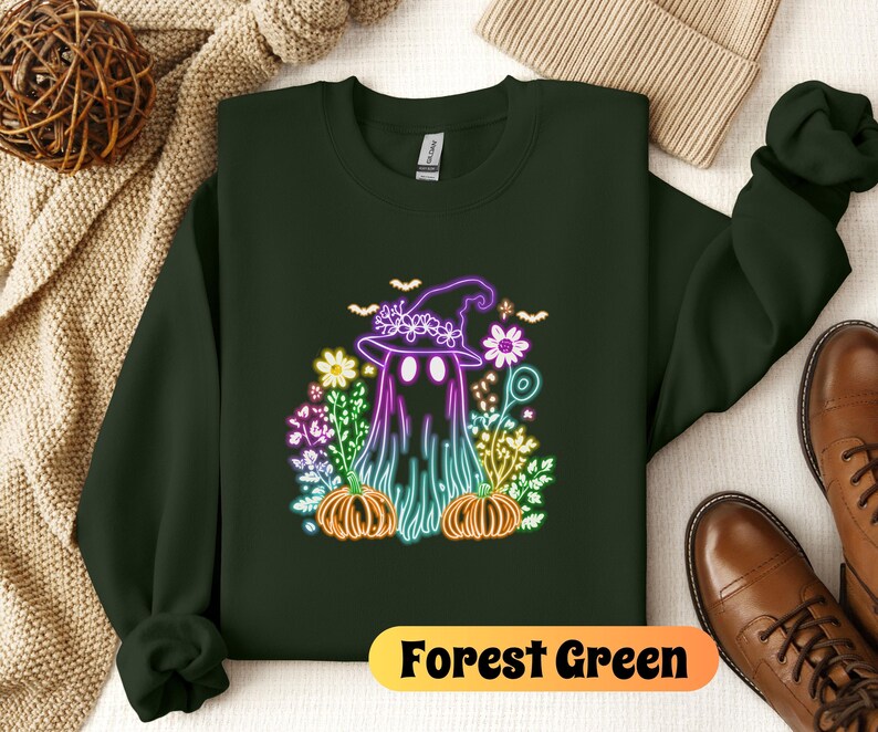 K&ouml;nnte beinhalten: Waldgr&uuml;ner Sweatshirt mit einem neongr&uuml;nen, violetten und rosafarbenen Geist, der einen Hexenhut tr&auml;gt und von Blumen und K&uuml;rbissen umgeben ist. Der Text "Forest Green" ist unten auf dem Bild aufgedruckt.