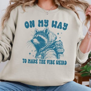 May include: A light beige sweatshirt with a blue graphic of a raccoon wearing a hooded jacket and a backpack. The text "On my way to make the vibe weird" is printed in blue below the raccoon.