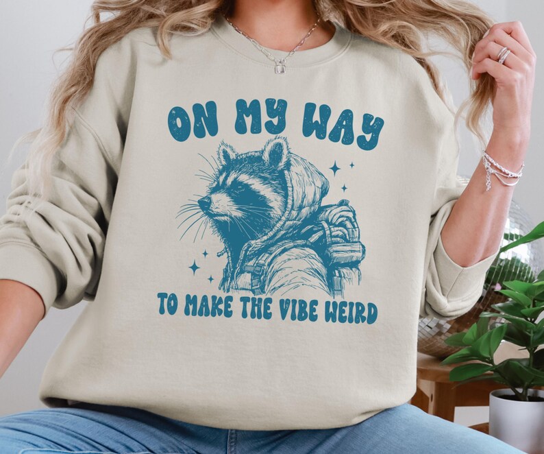 May include: A light beige sweatshirt with a blue graphic of a raccoon wearing a hooded jacket and a backpack. The text "On my way to make the vibe weird" is printed in blue below the raccoon.