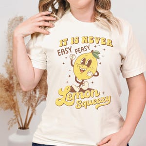 May include: A white t-shirt with a retro-style graphic of a smiling lemon. The text on the shirt reads "It is never easy peasy Lemon Squeezy".