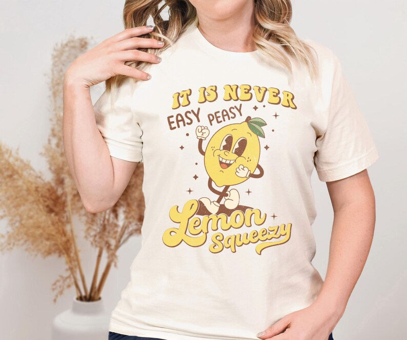 May include: A white t-shirt with a retro-style graphic of a smiling lemon. The text on the shirt reads "It is never easy peasy Lemon Squeezy".
