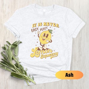May include: A white t-shirt with a yellow lemon graphic and the text "It is never easy peasy Lemon Squeezy". The shirt is available in the color Ash.