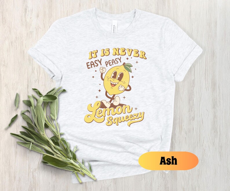 May include: A white t-shirt with a yellow lemon graphic and the text "It is never easy peasy Lemon Squeezy". The shirt is available in the color Ash.