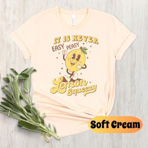 May include: A soft cream colored t-shirt with a retro cartoon lemon graphic and the text "It is never easy peasy Lemon Squeezy".