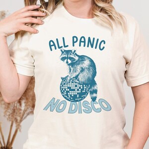 May include: A white t-shirt with a blue raccoon sitting on a disco ball. The text "All Panic No Disco" is printed on the shirt.