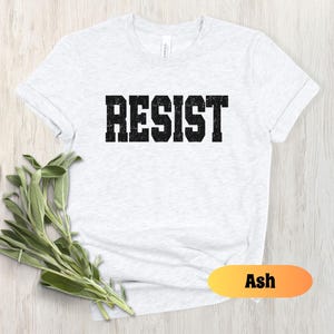 May include: Pale grey t-shirt featuring the word "RESIST" in large, black, block letters. The shirt has short sleeves and is made of a soft fabric. A sprig of green leaves is placed near the shirt. The word "Ash" is in a yellow and orange oval.