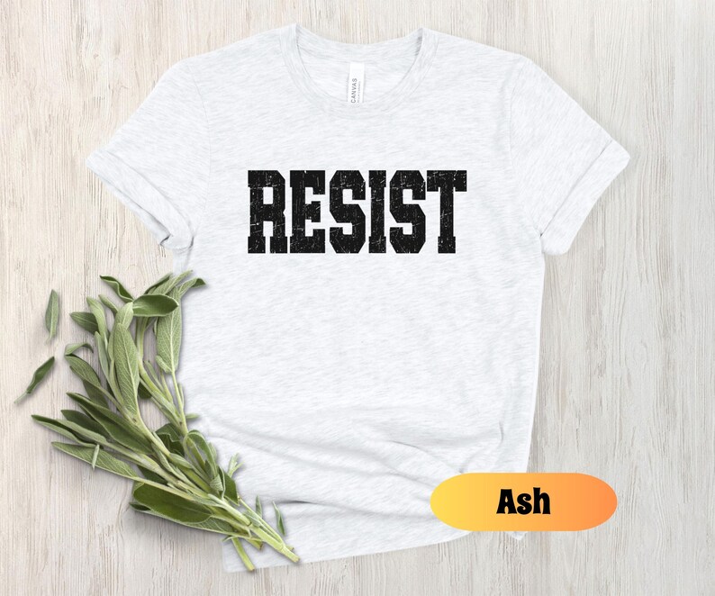 May include: Pale grey t-shirt featuring the word "RESIST" in large, black, block letters. The shirt has short sleeves and is made of a soft fabric. A sprig of green leaves is placed near the shirt. The word "Ash" is in a yellow and orange oval.