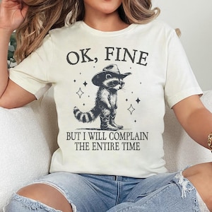 May include: A white t-shirt with a black and white illustration of a raccoon wearing a cowboy hat. The text "OK, FINE BUT I WILL COMPLAIN THE ENTIRE TIME" is printed on the shirt.