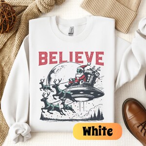 May include: White sweatshirt with a Christmas-themed graphic of Santa Claus in a spaceship with reindeer flying behind him. The text "BELIEVE" is printed above the graphic.