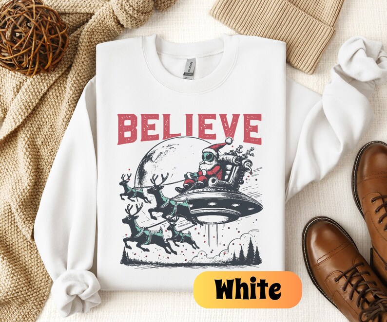 May include: White sweatshirt with a Christmas-themed graphic of Santa Claus in a spaceship with reindeer flying behind him. The text "BELIEVE" is printed above the graphic.