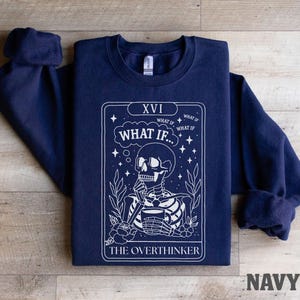 Puede incluir: Sudadera azul marino con un dise&ntilde;o de carta del tarot en blanco. El gr&aacute;fico presenta un esqueleto sosteniendo una taza, con el texto "WHAT IF..." y "THE OVERTHINKER". La sudadera tiene cuello redondo y mangas largas.