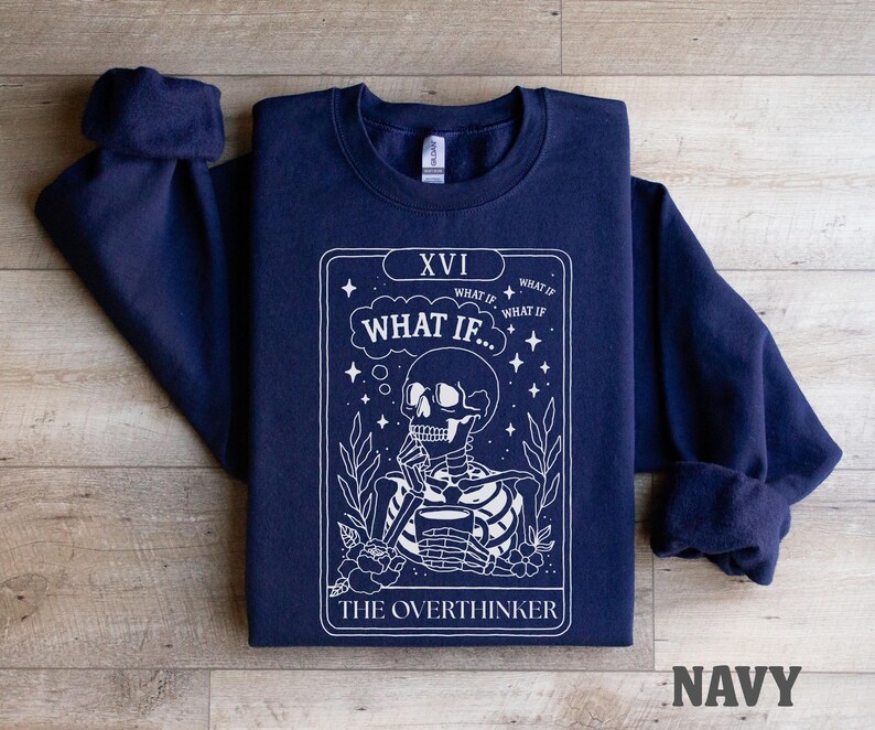 Puede incluir: Sudadera azul marino con un dise&ntilde;o de carta del tarot en blanco. El gr&aacute;fico presenta un esqueleto sosteniendo una taza, con el texto "WHAT IF..." y "THE OVERTHINKER". La sudadera tiene cuello redondo y mangas largas.