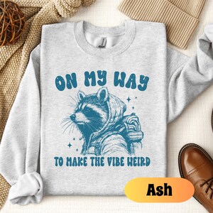 May include: A gray sweatshirt with a raccoon wearing a hooded jacket and the text "On my way to make the vibe weird". The sweatshirt is available in the color Ash.