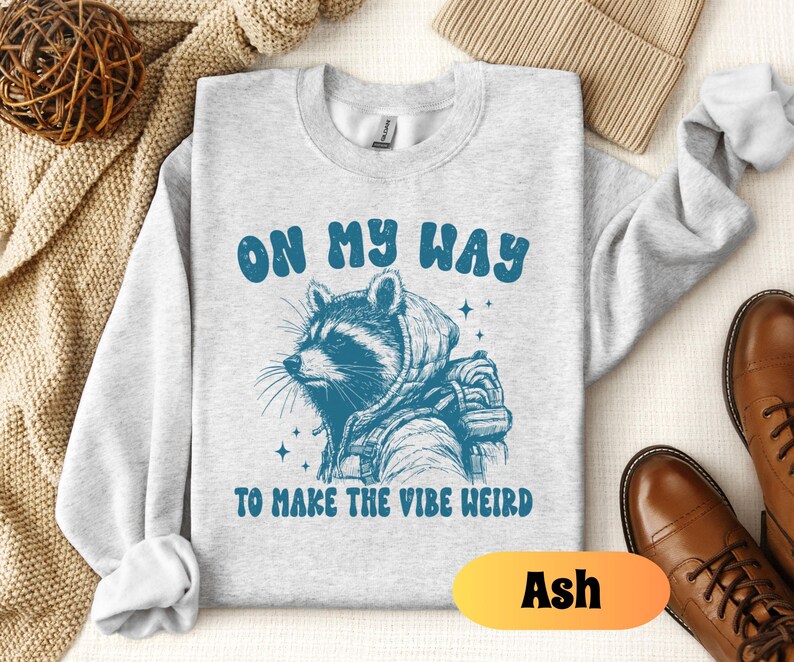 May include: A gray sweatshirt with a raccoon wearing a hooded jacket and the text "On my way to make the vibe weird". The sweatshirt is available in the color Ash.