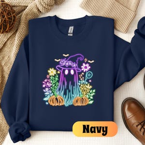 K&ouml;nnte beinhalten: Ein marineblauer Sweatshirt mit einer neonrosa, violetten und blauen Grafik eines Geistes, der einen Hexenhut tr&auml;gt und von Blumen und K&uuml;rbissen umgeben ist.