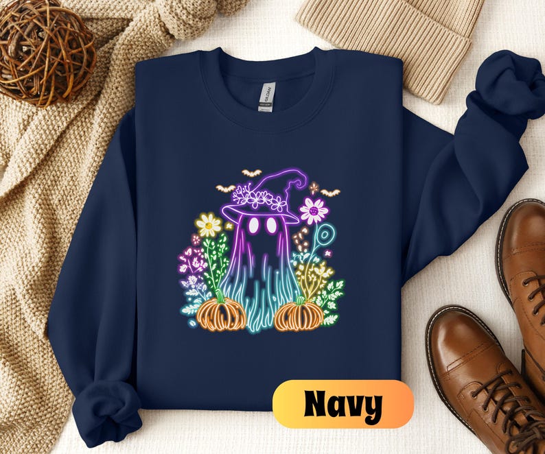 K&ouml;nnte beinhalten: Ein marineblauer Sweatshirt mit einer neonrosa, violetten und blauen Grafik eines Geistes, der einen Hexenhut tr&auml;gt und von Blumen und K&uuml;rbissen umgeben ist.