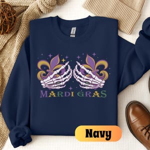 May include: Navy blue sweatshirt with a Mardi Gras design featuring two skeletal hands holding purple, green, and gold fleur-de-lis. The text "Mardi Gras" is printed below the hands.