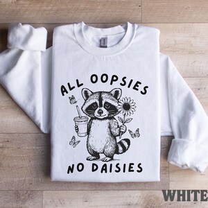 May include: White sweatshirt featuring a black and white illustration of a raccoon holding a drink and a sunflower, with butterflies. The text "ALL OOPSIES NO DAISIES" is printed on the front. The word "WHITE" is printed at the bottom.