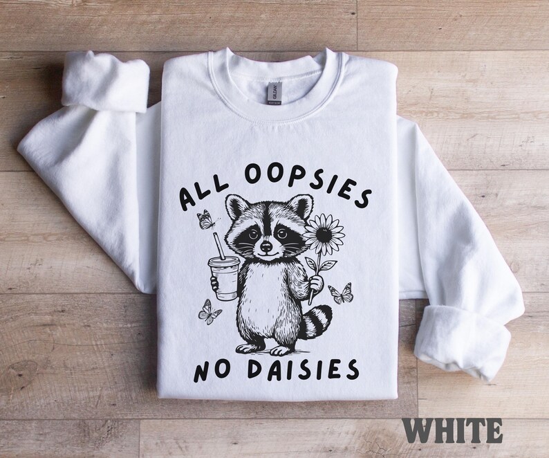 May include: White sweatshirt featuring a black and white illustration of a raccoon holding a drink and a sunflower, with butterflies. The text "ALL OOPSIES NO DAISIES" is printed on the front. The word "WHITE" is printed at the bottom.