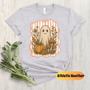 May include: Athletic Heather t-shirt featuring a whimsical Halloween design. The graphic includes a friendly ghost, pumpkin, mushrooms, and fall foliage within a striped frame. The shirt is light gray.
