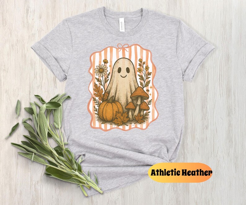 May include: Athletic Heather t-shirt featuring a whimsical Halloween design. The graphic includes a friendly ghost, pumpkin, mushrooms, and fall foliage within a striped frame. The shirt is light gray.