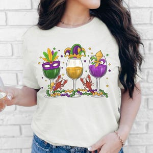 May include: A white t-shirt with a Mardi Gras themed design featuring three wine glasses with different colored liquids, a jester hat, a purple and green mask, and a fleur-de-lis. The glasses are surrounded by green and purple beads and stars.