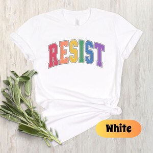 May include: White t-shirt with the word "RESIST" in rainbow colors. The word is in a varsity style font.