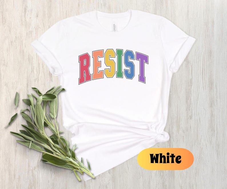 May include: White t-shirt with the word "RESIST" in rainbow colors. The word is in a varsity style font.