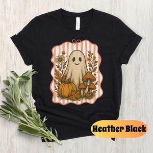 May include: Heather black t-shirt featuring a whimsical Halloween design. The graphic includes a friendly ghost, pumpkin, mushrooms, and fall foliage within a striped frame. The shirt is made of soft material.