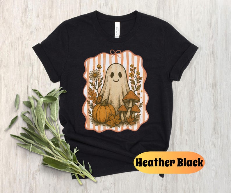 May include: Heather black t-shirt featuring a whimsical Halloween design. The graphic includes a friendly ghost, pumpkin, mushrooms, and fall foliage within a striped frame. The shirt is made of soft material.