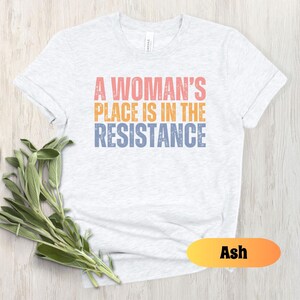 May include: Ash-colored t-shirt with the text "A Woman's Place Is In The Resistance" in coral, orange, and blue. The shirt is displayed on a light wood surface with a sprig of green leaves.