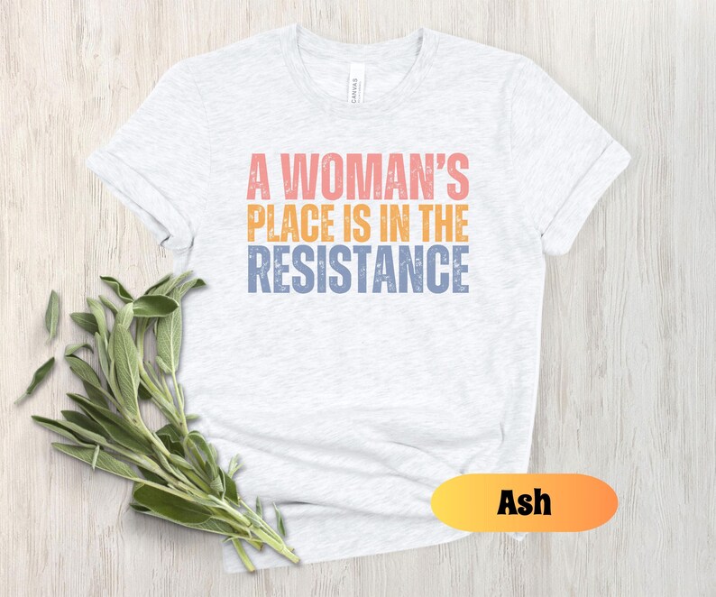 May include: Ash-colored t-shirt with the text "A Woman's Place Is In The Resistance" in coral, orange, and blue. The shirt is displayed on a light wood surface with a sprig of green leaves.